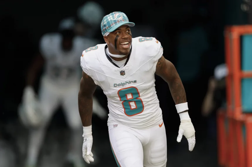 Jevon Holland in weißer Uniform der Miami Dolphins und blaukarierter Kappe, identifiziert als Jevon Holland, lächelt breit beim Laufen. Er trägt ein Trikot mit der Nummer 8. Im Hintergrund ist ein dunkler Tunnel zu sehen, in dessen Schatten ein anderer Spieler leicht zu erkennen ist. Diese Beschreibung wurde mit der FootballR KI automatisch generiert.