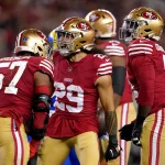 Dre Greenlaw und Talanoa Hufanga - Drei Footballspieler in rot-goldenen Uniformen und Helmen stehen auf einem Feld. Der Spieler in der Mitte, Nummer 29, wirkt aufgeregt und hat die Arme leicht erhoben. Zwei weitere Spieler, Nummer 57 und ein weiterer mit Helm, stehen dicht vor ihm und verkörpern den Geist der 49ers-Stars. Im Hintergrund ist eine verschwommene Menschenmenge zu sehen. Diese Beschreibung wurde mit der FootballR KI automatisch generiert.