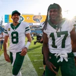 Aaron Rodgers und Davante Adams in grün-weißen Trikots laufen lächelnd über ein Fußballfeld. Einer trägt ein Trikot mit der Nummer 8, der andere die Nummer 17. Im Hintergrund zeigt eine Anzeigetafel die Mannschaften und den Punktestand. Auf den Tribünen sitzen Zuschauer, und die Sonne geht unter, was für ein warmes Leuchten sorgt. Diese Beschreibung wurde mit der FootballR KI automatisch generiert.