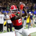 Nitro Tuggle in rot-schwarzer Uniform mit der Nummer 2 fängt während des Spiels einen Ball. Sein roter Helm trägt ein Tuggle-Logo mit schwarzem Streifen. Auf dem Spielfeld wimmelt es von Zuschauern und Fotografen, während das Teampersonal im Hintergrund zusieht. Diese Beschreibung wurde mit der FootballR KI automatisch generiert.