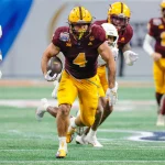 Cam Skattebo, in einem kastanienbraunen Trikot mit der Nummer 4 und gelben Hosen, stürmt mit dem Football in der Hand über den Rasen. Umgeben von Teamkollegen und Gegnern in weiß-orangenen Uniformen bleibt er konzentriert und verkörpert die Intensität dieses dynamischen Spielmoments. Diese Beschreibung wurde mit der FootballR KI automatisch generiert.