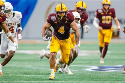 Cam Skattebo, in einem kastanienbraunen Trikot mit der Nummer 4 und gelben Hosen, stürmt mit dem Football in der Hand über den Rasen. Umgeben von Teamkollegen und Gegnern in weiß-orangenen Uniformen bleibt er konzentriert und verkörpert die Intensität dieses dynamischen Spielmoments. Diese Beschreibung wurde mit der FootballR KI automatisch generiert.