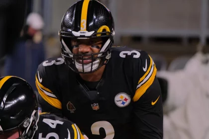 Quarterback Russell Wilson in schwarz-goldener Uniform, der Russell Wilson ähnelt, trägt die Nummer 3 und bereitet sich konzentriert auf dem Spielfeld vor. Schwarze Augenstreifen betonen seinen entschlossenen Blick. In der Nähe ist ein weiterer Spieler mit der Nummer 54 vor dem unscharfen Hintergrund teilweise zu erkennen, was an ein Spiel der New York Giants erinnert. Diese Beschreibung wurde mit der FootballR KI automatisch generiert.