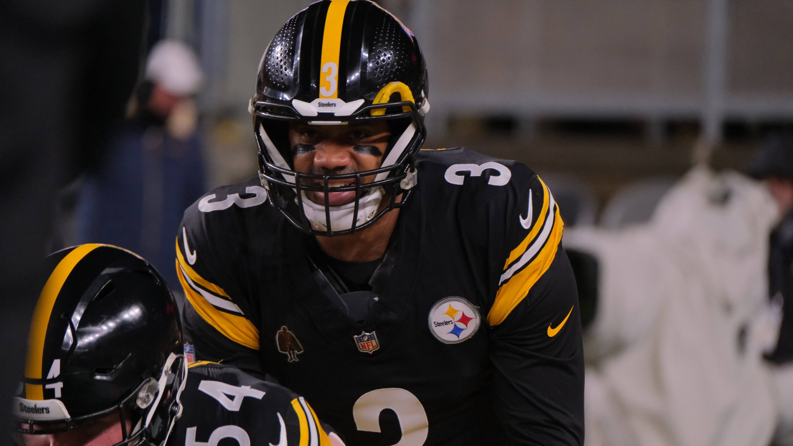 Quarterback Russell Wilson in schwarz-goldener Uniform, der Russell Wilson ähnelt, trägt die Nummer 3 und bereitet sich konzentriert auf dem Spielfeld vor. Schwarze Augenstreifen betonen seinen entschlossenen Blick. In der Nähe ist ein weiterer Spieler mit der Nummer 54 vor dem unscharfen Hintergrund teilweise zu erkennen, was an ein Spiel der New York Giants erinnert. Diese Beschreibung wurde mit der FootballR KI automatisch generiert.