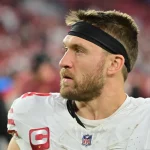 Ein Footballspieler, der Kyle Juszczyk ähnelt, trägt auf dem Spielfeld ein weißes Trikot mit roten Akzenten und einem NFL-Logo am Kragen. Er hat ein schwarzes Stirnband, einen Bart und trägt ein Kapitänsabzeichen. Hinter ihm sind Fans zu sehen und das leuchtende Rot des Stadions. Diese Beschreibung wurde mit der FootballR KI automatisch generiert.