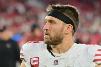 Ein Footballspieler, der Kyle Juszczyk ähnelt, trägt auf dem Spielfeld ein weißes Trikot mit roten Akzenten und einem NFL-Logo am Kragen. Er hat ein schwarzes Stirnband, einen Bart und trägt ein Kapitänsabzeichen. Hinter ihm sind Fans zu sehen und das leuchtende Rot des Stadions. Diese Beschreibung wurde mit der FootballR KI automatisch generiert.