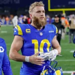 Cooper Kupp in blau-gelber „Rams“-Uniform mit der Nummer 10 läuft mit Helm über das Spielfeld. Er hat einen Bart und kurze blonde Haare. Neben ihm steht eine weitere Person, die ein blaues „LAFD“-Sweatshirt und eine Mütze trägt. Im Hintergrund sind ein Footballfeld und Menschen zu sehen. Diese Beschreibung wurde mit der FootballR KI automatisch generiert.