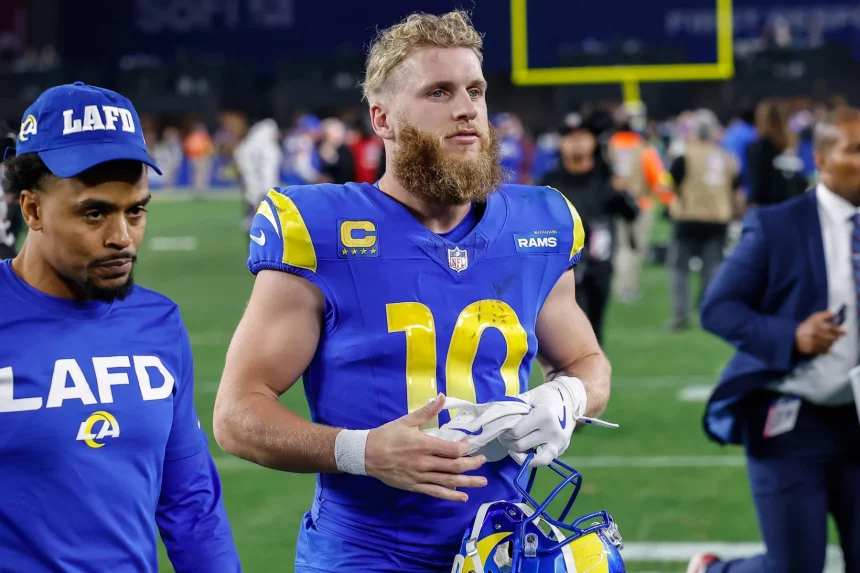 Cooper Kupp in blau-gelber „Rams“-Uniform mit der Nummer 10 läuft mit Helm über das Spielfeld. Er hat einen Bart und kurze blonde Haare. Neben ihm steht eine weitere Person, die ein blaues „LAFD“-Sweatshirt und eine Mütze trägt. Im Hintergrund sind ein Footballfeld und Menschen zu sehen. Diese Beschreibung wurde mit der FootballR KI automatisch generiert.