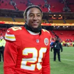Justin Reid in der roten Uniform der Kansas City Chiefs, Nummer 20, lächelt auf dem Spielfeld. Das Stadion ist fast leer, im Hintergrund stehen ein paar Leute. Justin Reid hat kurze Haare und trägt einen schwarzen Halswärmer. Die Atmosphäre lässt vermuten, dass ein Spiel gerade zu Ende gegangen ist. Diese Beschreibung wurde mit der FootballR KI automatisch generiert.