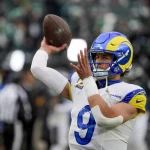 NFC West - Matthew Stafford im Trikot der Los Angeles Rams, mit einem blau-weißen Helm und einem gelben Widderhorn-Design, bereitet sich auf einen Footballwurf vor. Die Trikotnummer 9 weist auf Matthew Stafford hin. Ziel des Passes ist Davante Adams. Im Hintergrund ist ein Freiluftstadion mit verschwommenen Zuschauern zu sehen. Diese Beschreibung wurde mit der FootballR KI automatisch generiert.