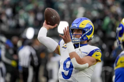 NFC West - Matthew Stafford im Trikot der Los Angeles Rams, mit einem blau-weißen Helm und einem gelben Widderhorn-Design, bereitet sich auf einen Footballwurf vor. Die Trikotnummer 9 weist auf Matthew Stafford hin. Ziel des Passes ist Davante Adams. Im Hintergrund ist ein Freiluftstadion mit verschwommenen Zuschauern zu sehen. Diese Beschreibung wurde mit der FootballR KI automatisch generiert.