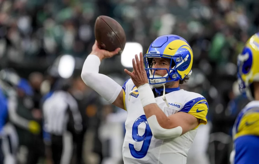 NFC West - Matthew Stafford im Trikot der Los Angeles Rams, mit einem blau-weißen Helm und einem gelben Widderhorn-Design, bereitet sich auf einen Footballwurf vor. Die Trikotnummer 9 weist auf Matthew Stafford hin. Ziel des Passes ist Davante Adams. Im Hintergrund ist ein Freiluftstadion mit verschwommenen Zuschauern zu sehen. Diese Beschreibung wurde mit der FootballR KI automatisch generiert.