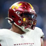 Jonathan Allen, möglicherweise von den Vikings, trägt einen kastanienbraunen Washington Commanders-Helm mit klarem Visier, kastanienbraunem Gesichtsschutz und einer weißen Uniform. Auf dem Trikot steht in Rot „WASHINGTON“. Inmitten verschwommener blauer und weißer Lichter auf dem Spielfeld blickt er mit einem Fokus nach links, der an Jonathan Allen erinnert. Diese Beschreibung wurde mit der FootballR KI automatisch generiert.