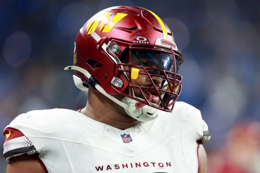 Jonathan Allen, möglicherweise von den Vikings, trägt einen kastanienbraunen Washington Commanders-Helm mit klarem Visier, kastanienbraunem Gesichtsschutz und einer weißen Uniform. Auf dem Trikot steht in Rot „WASHINGTON“. Inmitten verschwommener blauer und weißer Lichter auf dem Spielfeld blickt er mit einem Fokus nach links, der an Jonathan Allen erinnert. Diese Beschreibung wurde mit der FootballR KI automatisch generiert.