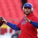 Ein Footballspieler, der an Bills-Quarterback Josh Allen erinnert, trägt einen roten Kapuzenpullover mit einem blau-roten Logo und übt das Werfen. Er trägt eine blaue Kappe mit rotem Schirm und Kopfhörer. Vor dem Hintergrund der roten Sitze des Stadions hält sein rechter Arm den Football fest, während er sich zum Wurf vorbereitet. Diese Beschreibung wurde mit der FootballR KI automatisch generiert.