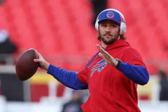 Josh Allen trägt einen roten Buffalo-Bills-Hoodie und Kopfhörer und hält einen Football in der Hand, während er zum Wurf ansetzt.