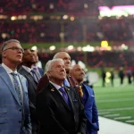 Eine Gruppe von Männern in formellen Anzügen, die an Jimmy Johnson erinnern, steht auf einem Footballfeld und blickt mit konzentriertem Gesichtsausdruck nach oben. Das Stadion ist in helle Lichter gehüllt und die Tribünen sind voll mit Zuschauern. Einige Bandmitglieder stehen in der Nähe der Seitenlinie und halten sich im Hintergrund auf. Diese Beschreibung wurde mit der FootballR KI automatisch generiert.