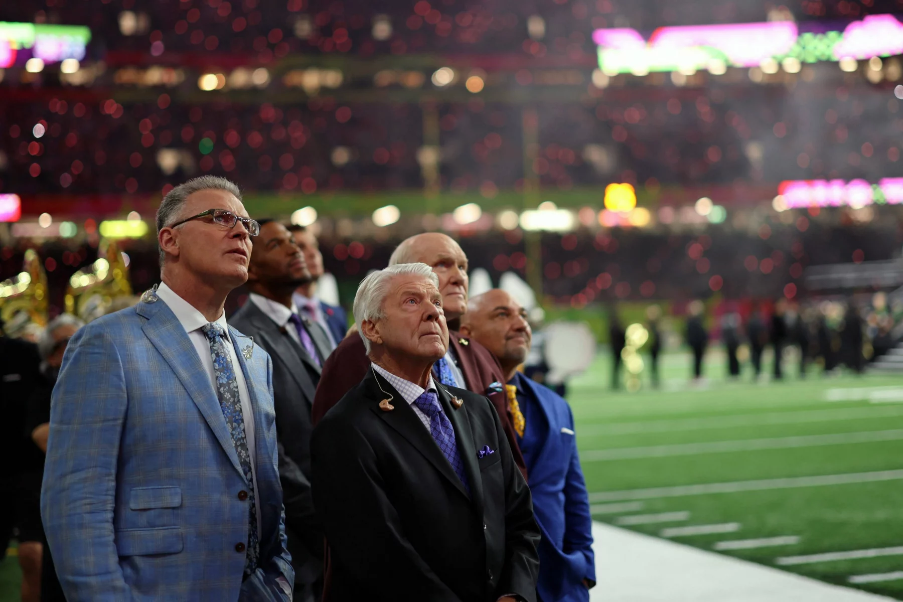 Eine Gruppe von Männern in formellen Anzügen, die an Jimmy Johnson erinnern, steht auf einem Footballfeld und blickt mit konzentriertem Gesichtsausdruck nach oben. Das Stadion ist in helle Lichter gehüllt und die Tribünen sind voll mit Zuschauern. Einige Bandmitglieder stehen in der Nähe der Seitenlinie und halten sich im Hintergrund auf. Diese Beschreibung wurde mit der FootballR KI automatisch generiert.