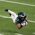 Zack Baun mit der Nummer 53 taucht in einer dunkelgrünen Uniform horizontal ab und hält einen Football in der Hand. Es ist der agile Zack Baun, der einen passenden Helm und weiße Hosen mit schwarzen Stollen trägt. Das Feld ist mit weißen Yard-Linien übersät und fängt die Intensität einer Indoor-Stadionszene ein. Diese Beschreibung wurde mit der FootballR KI automatisch generiert.