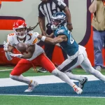 DeAndre Hopkins in rot-weißer Uniform fängt einen Ball in der Nähe der Endzone, dicht gefolgt von einem blaugrün gekleideten Verteidiger. Die actionreiche Szene erinnert an DeAndre Hopkins, der einen seiner der Schwerkraft trotzenden Fänge versucht, während Schiedsrichter und Medienvertreter im Hintergrund zuschauen. Diese Beschreibung wurde mit der FootballR KI automatisch generiert.