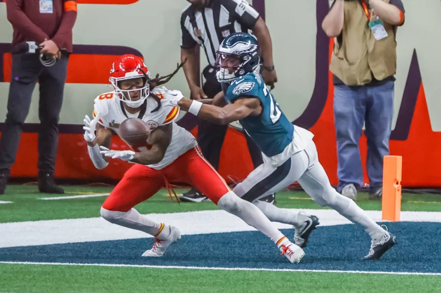 DeAndre Hopkins in rot-weißer Uniform fängt einen Ball in der Nähe der Endzone, dicht gefolgt von einem blaugrün gekleideten Verteidiger. Die actionreiche Szene erinnert an DeAndre Hopkins, der einen seiner der Schwerkraft trotzenden Fänge versucht, während Schiedsrichter und Medienvertreter im Hintergrund zuschauen. Diese Beschreibung wurde mit der FootballR KI automatisch generiert.