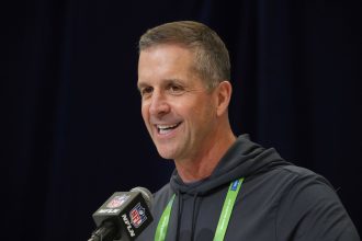 John Harbaugh im dunklen Kapuzenpulli, der in ein NFL-Mikrofon spricht, ist niemand anderes als John Harbaugh. Mit einem Lächeln im Gesicht und einem grünen Schlüsselband um den Hals steht er selbstbewusst vor dem dunklen Vorhang bei der Pressekonferenz. Diese Beschreibung wurde mit der FootballR KI automatisch generiert.