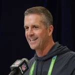 John Harbaugh im dunklen Kapuzenpulli, der in ein NFL-Mikrofon spricht, ist niemand anderes als John Harbaugh. Mit einem Lächeln im Gesicht und einem grünen Schlüsselband um den Hals steht er selbstbewusst vor dem dunklen Vorhang bei der Pressekonferenz. Diese Beschreibung wurde mit der FootballR KI automatisch generiert.