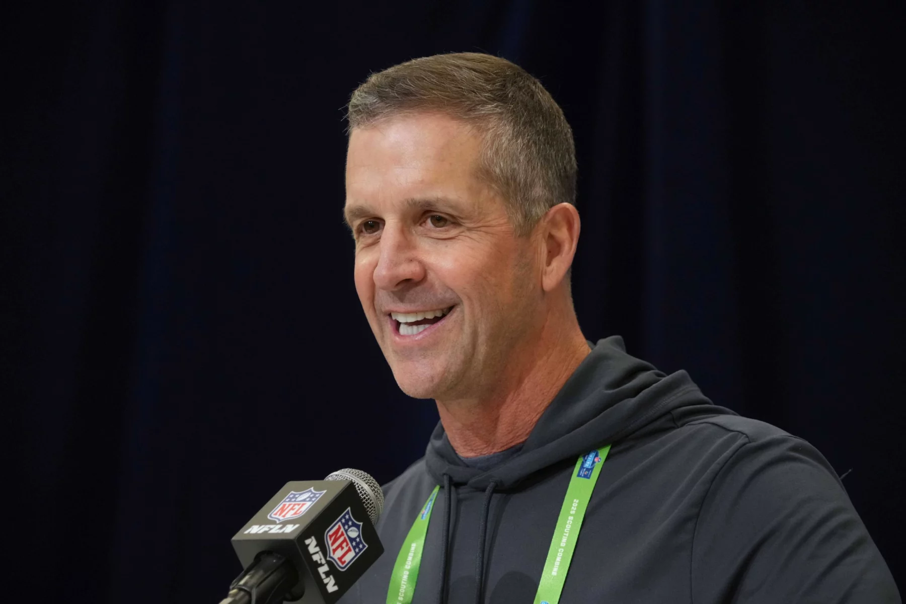 John Harbaugh im dunklen Kapuzenpulli, der in ein NFL-Mikrofon spricht, ist niemand anderes als John Harbaugh. Mit einem Lächeln im Gesicht und einem grünen Schlüsselband um den Hals steht er selbstbewusst vor dem dunklen Vorhang bei der Pressekonferenz. Diese Beschreibung wurde mit der FootballR KI automatisch generiert.