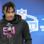 Kevin Winston Jr., mit geflochtenen Haaren und einem schwarzen New Era Hoodie mit der Nummer 54 und dem NFL-Logo, steht selbstbewusst am Mikrofon. Hinter ihm ragt die legendäre Kulisse des NFL Combine auf, die sowohl das NFL-Emblem als auch Microsoft Surface-Schilder zeigt und so die perfekte Kulisse für dieses Presseevent bildet. Diese Beschreibung wurde mit der FootballR KI automatisch generiert.