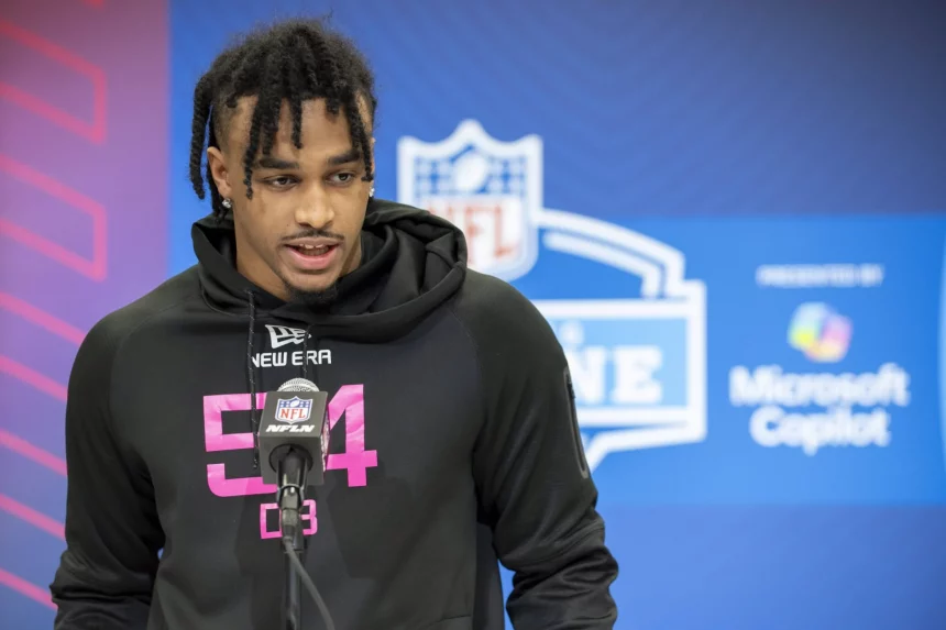 Kevin Winston Jr., mit geflochtenen Haaren und einem schwarzen New Era Hoodie mit der Nummer 54 und dem NFL-Logo, steht selbstbewusst am Mikrofon. Hinter ihm ragt die legendäre Kulisse des NFL Combine auf, die sowohl das NFL-Emblem als auch Microsoft Surface-Schilder zeigt und so die perfekte Kulisse für dieses Presseevent bildet. Diese Beschreibung wurde mit der FootballR KI automatisch generiert.