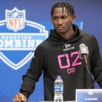 Zy Alexander, ein Footballspieler, steht während des NFL Scouting Combine am Podium. Er trägt einen schwarzen Kapuzenpullover mit der Aufschrift „02 DB“ in Pink und hat ordentlich geflochtene Haare. Vor ihm stehen ein Mikrofon und eine Wasserflasche, während das NFL-Logo und der Schriftzug „Scouting Combine“ den blauen Hintergrund zieren. Diese Beschreibung wurde mit der FootballR KI automatisch generiert.