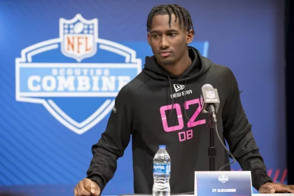 Zy Alexander, ein Footballspieler, steht während des NFL Scouting Combine am Podium. Er trägt einen schwarzen Kapuzenpullover mit der Aufschrift „02 DB“ in Pink und hat ordentlich geflochtene Haare. Vor ihm stehen ein Mikrofon und eine Wasserflasche, während das NFL-Logo und der Schriftzug „Scouting Combine“ den blauen Hintergrund zieren. Diese Beschreibung wurde mit der FootballR KI automatisch generiert.