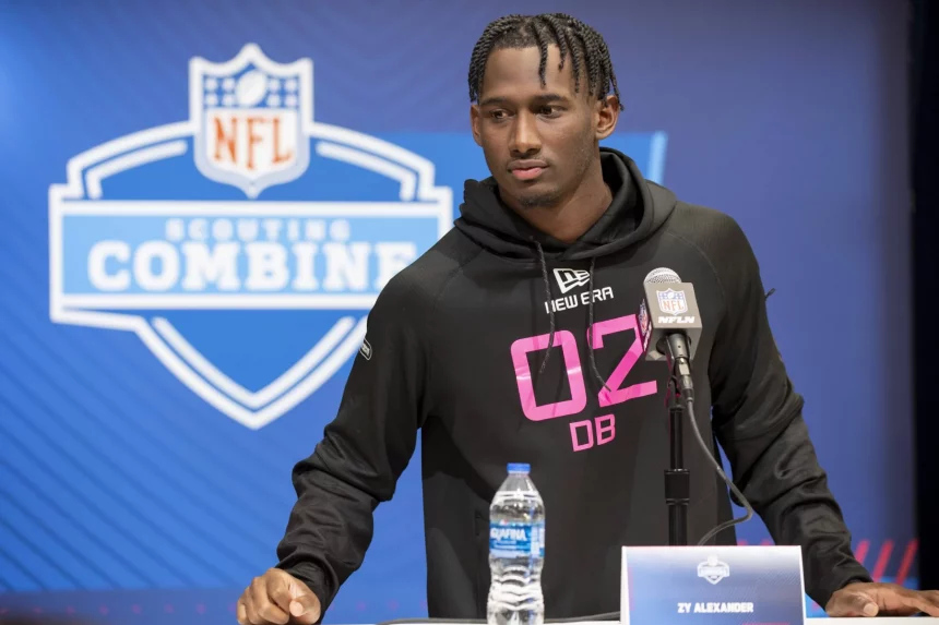 Zy Alexander, ein Footballspieler, steht während des NFL Scouting Combine am Podium. Er trägt einen schwarzen Kapuzenpullover mit der Aufschrift „02 DB“ in Pink und hat ordentlich geflochtene Haare. Vor ihm stehen ein Mikrofon und eine Wasserflasche, während das NFL-Logo und der Schriftzug „Scouting Combine“ den blauen Hintergrund zieren. Diese Beschreibung wurde mit der FootballR KI automatisch generiert.