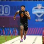 Beim NFL Combine sprintet Matthew Golden in einem schwarzen ärmellosen Oberteil mit der Aufschrift „16“ und „WO“ die Strecke hinunter. Über ihm hängt ein blaues Banner mit der Aufschrift „40 YARD“, während rote Zeitsensoren den Weg säumen. Im Hintergrund entfaltet sich inmitten dieser aufregenden Darbietung sportlicher Leistungsfähigkeit dezent ein Logo. Diese Beschreibung wurde mit der FootballR KI automatisch generiert.