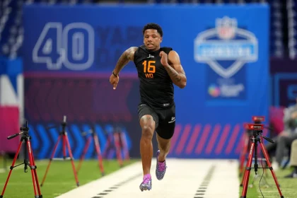 Beim NFL Combine sprintet Matthew Golden in einem schwarzen ärmellosen Oberteil mit der Aufschrift „16“ und „WO“ die Strecke hinunter. Über ihm hängt ein blaues Banner mit der Aufschrift „40 YARD“, während rote Zeitsensoren den Weg säumen. Im Hintergrund entfaltet sich inmitten dieser aufregenden Darbietung sportlicher Leistungsfähigkeit dezent ein Logo. Diese Beschreibung wurde mit der FootballR KI automatisch generiert.