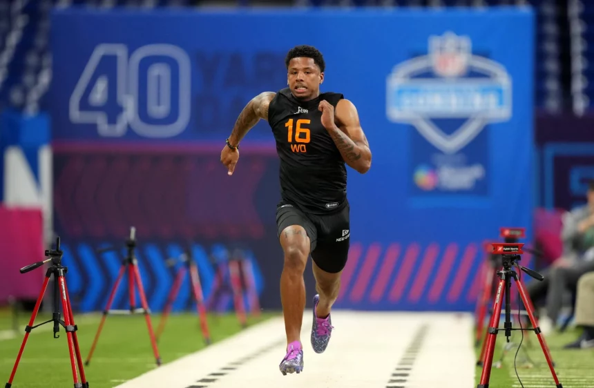 Beim NFL Combine sprintet Matthew Golden in einem schwarzen ärmellosen Oberteil mit der Aufschrift „16“ und „WO“ die Strecke hinunter. Über ihm hängt ein blaues Banner mit der Aufschrift „40 YARD“, während rote Zeitsensoren den Weg säumen. Im Hintergrund entfaltet sich inmitten dieser aufregenden Darbietung sportlicher Leistungsfähigkeit dezent ein Logo. Diese Beschreibung wurde mit der FootballR KI automatisch generiert.