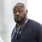 Mike Tomlin mit Bart und Schnurrbart trägt drinnen ein marineblaues Nike-Sweatshirt. Um seinen Hals hängt ein Notre-Dame-Schlüsselband, während er einen ernsten Gesichtsausdruck behält. Der verschwommene, helle Hintergrund lenkt den Fokus auf sein gelassenes Auftreten. Diese Beschreibung wurde mit der FootballR KI automatisch generiert.