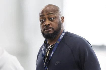 Mike Tomlin mit Bart und Schnurrbart trägt drinnen ein marineblaues Nike-Sweatshirt. Um seinen Hals hängt ein Notre-Dame-Schlüsselband, während er einen ernsten Gesichtsausdruck behält. Der verschwommene, helle Hintergrund lenkt den Fokus auf sein gelassenes Auftreten. Diese Beschreibung wurde mit der FootballR KI automatisch generiert.