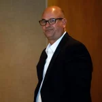 Jeremy Pruitt, rasiert und mit Brille, steht in einem Raum. Er trägt ein schwarzes Jackett über einem weißen Hemd. Sein Gesichtsausdruck ist neutral, als er in die Kamera blickt. Im Hintergrund sind eine braune Wand und eine Holzvertäfelung zu sehen, die der Szenerie Tiefe verleihen. Diese Beschreibung wurde mit der FootballR KI automatisch generiert.