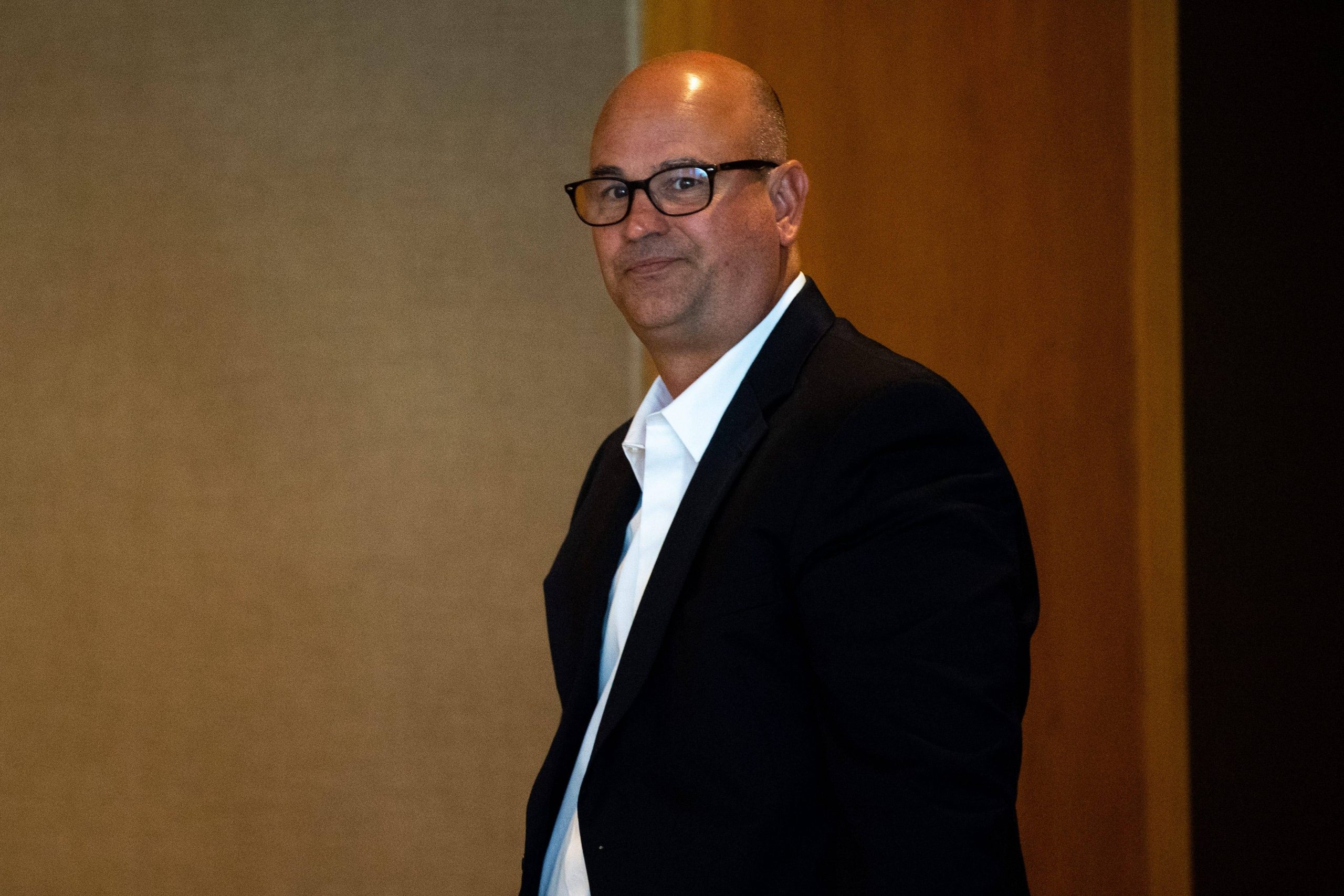 Jeremy Pruitt, rasiert und mit Brille, steht in einem Raum. Er trägt ein schwarzes Jackett über einem weißen Hemd. Sein Gesichtsausdruck ist neutral, als er in die Kamera blickt. Im Hintergrund sind eine braune Wand und eine Holzvertäfelung zu sehen, die der Szenerie Tiefe verleihen. Diese Beschreibung wurde mit der FootballR KI automatisch generiert.