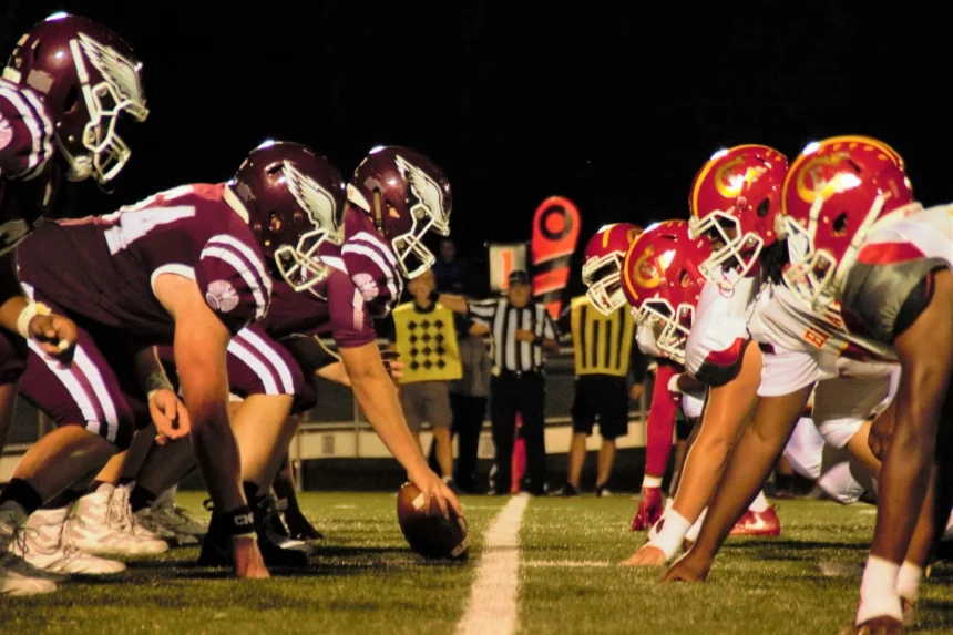 Unter den hellen Stadionlichtern eines klassischen College-Football-Duells stehen sich zwei amerikanische Teams an der Line of Scrimmage gegenüber. Die kastanienbraun gekleideten Spieler treten gegen ihre Rivalen in Rot und Gelb an. Schiedsrichter und eine Yard-Markierung wachen auf dem sattgrünen Rasen und präsentieren die aufstrebenden Talente der USA. Diese Beschreibung wurde mit der FootballR KI automatisch generiert.