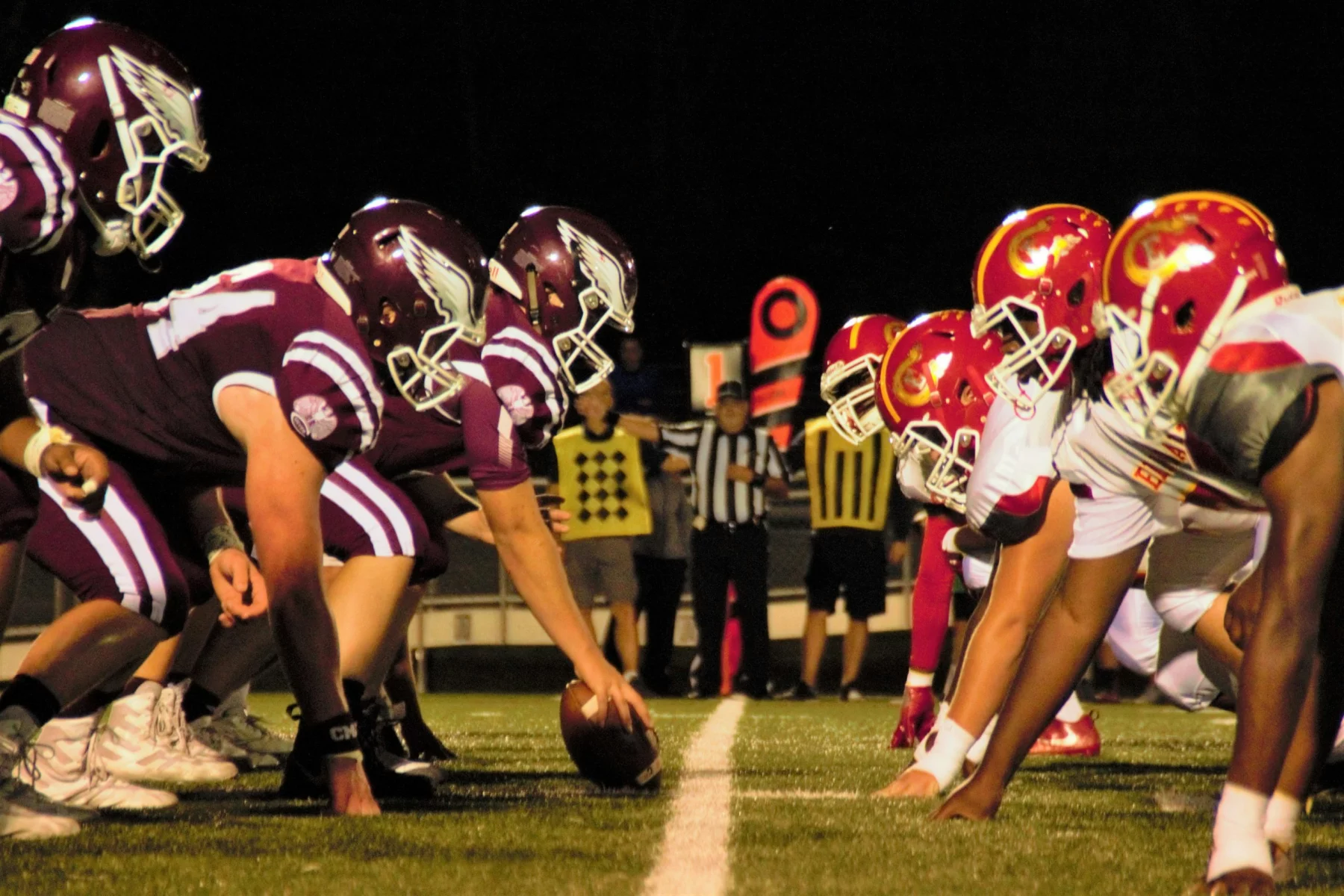Unter den hellen Stadionlichtern eines klassischen College-Football-Duells stehen sich zwei amerikanische Teams an der Line of Scrimmage gegenüber. Die kastanienbraun gekleideten Spieler treten gegen ihre Rivalen in Rot und Gelb an. Schiedsrichter und eine Yard-Markierung wachen auf dem sattgrünen Rasen und präsentieren die aufstrebenden Talente der USA. Diese Beschreibung wurde mit der FootballR KI automatisch generiert.