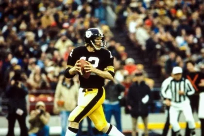 Ein Quarterback von Pittsburgh in schwarz-gelber Uniform bereitet sich während des Spiels auf einen Pass vor. Mit einem schwarzen Helm mit dem Teamlogo steht er inmitten jubelnder Zuschauer. Der Schiedsrichter beobachtet gespannt, wie sich auf dem leuchtend grünen Spielfeld der entscheidende Moment nähert. Diese Beschreibung wurde mit der FootballR KI automatisch generiert.