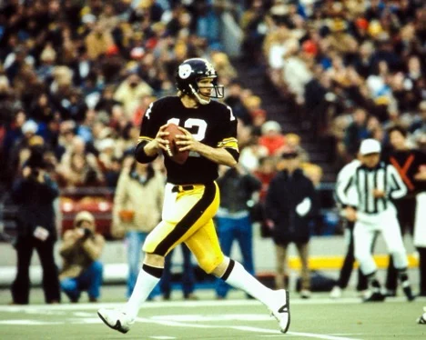 Ein Quarterback von Pittsburgh in schwarz-gelber Uniform bereitet sich während des Spiels auf einen Pass vor. Mit einem schwarzen Helm mit dem Teamlogo steht er inmitten jubelnder Zuschauer. Der Schiedsrichter beobachtet gespannt, wie sich auf dem leuchtend grünen Spielfeld der entscheidende Moment nähert. Diese Beschreibung wurde mit der FootballR KI automatisch generiert.