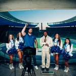 Fünf Personen stehen auf einem Podest im Berliner Olympiastadion: ein Mann im blauen Hemd mit erhobener Faust, ein anderer in Weiß mit einem Mikrofon und drei Cheerleader in Indianapolis-Colts-Uniformen mit Pompons. Lichttechnik und ein Kamerastativ sind zu sehen. Diese Beschreibung wurde mit der FootballR KI automatisch generiert.