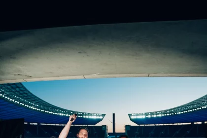 Fünf Personen stehen auf einem Podest im Berliner Olympiastadion: ein Mann im blauen Hemd mit erhobener Faust, ein anderer in Weiß mit einem Mikrofon und drei Cheerleader in Indianapolis-Colts-Uniformen mit Pompons. Lichttechnik und ein Kamerastativ sind zu sehen. Diese Beschreibung wurde mit der FootballR KI automatisch generiert.