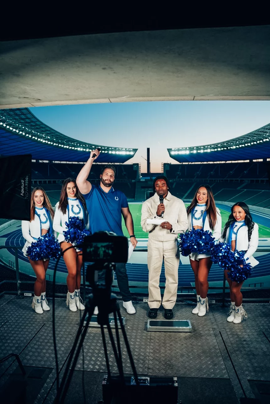 Fünf Personen stehen auf einem Podest im Berliner Olympiastadion: ein Mann im blauen Hemd mit erhobener Faust, ein anderer in Weiß mit einem Mikrofon und drei Cheerleader in Indianapolis-Colts-Uniformen mit Pompons. Lichttechnik und ein Kamerastativ sind zu sehen. Diese Beschreibung wurde mit der FootballR KI automatisch generiert.