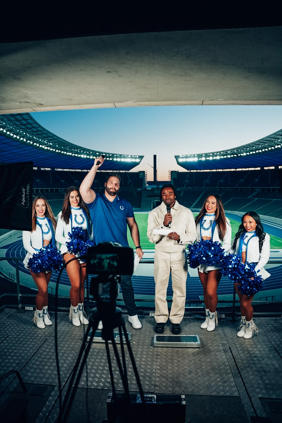 Fünf Personen stehen auf einem Podest im Berliner Olympiastadion: ein Mann im blauen Hemd mit erhobener Faust, ein anderer in Weiß mit einem Mikrofon und drei Cheerleader in Indianapolis-Colts-Uniformen mit Pompons. Lichttechnik und ein Kamerastativ sind zu sehen. Diese Beschreibung wurde mit der FootballR KI automatisch generiert.