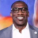 Shannon Sharpe mit schwarzer Brille, grauem Jackett und weißem Hemd steht vor einem bunten, unscharfen Hintergrund. Er hat kurze Haare, einen Spitzbart und eine weiße Blumenbrosche – eine Reminiszenz an Shannon Sharpes klassischen Stil. Diese Beschreibung wurde mit der FootballR KI automatisch generiert.