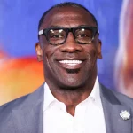 Shannon Sharpe mit schwarzer Brille, grauem Jackett und weißem Hemd steht vor einem bunten, unscharfen Hintergrund. Er hat kurze Haare, einen Spitzbart und eine weiße Blumenbrosche – eine Reminiszenz an Shannon Sharpes klassischen Stil. Diese Beschreibung wurde mit der FootballR KI automatisch generiert.